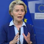 Een afbeelding van Ursula von der Leyen, voorzitter van de Europese Commissie, met op de achtergrond een vlag van de EU en een logo van de EU