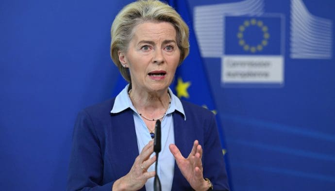 Een afbeelding van Ursula von der Leyen, voorzitter van de Europese Commissie, met op de achtergrond een vlag van de EU en een logo van de EU