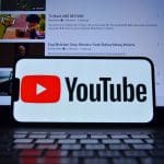 Het logo van Youtube op een mobiele telefoon met op de achtergrond een laptop met het YouTube interface en verschillende video's